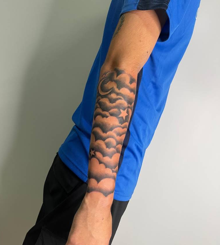 Brazo con tatuaje de nubes negras, luna creciente y estrellas en estilo sombreado.