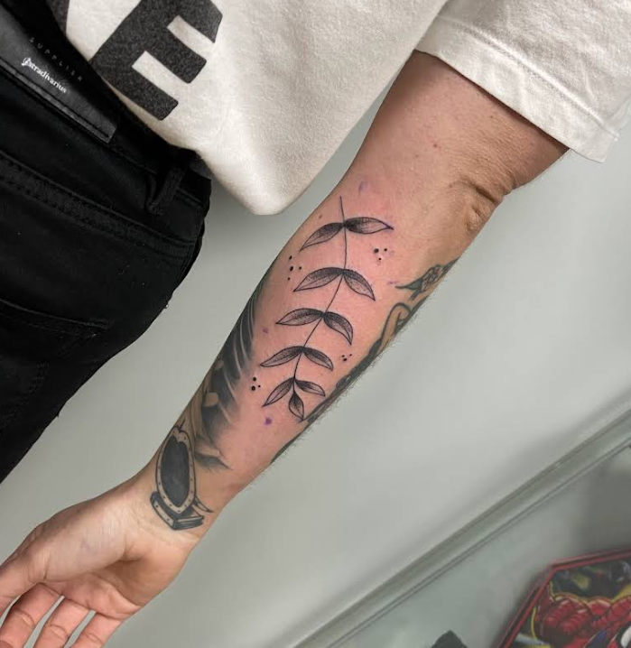 Tatuaje de rama con hojas y pequeños puntos, ubicado en el antebrazo.